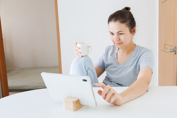Woman touching a tablet screen while holding a cup of coffee at home. Using touchscreen device, browsing online, internet technology and digital lifestyle. © soya