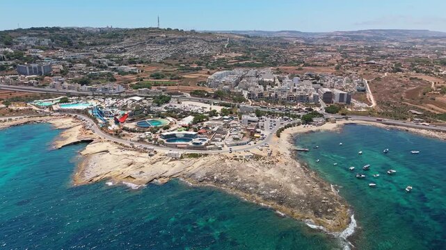 Baħar iċ-Ċagħaq, Malta aqua park with waterslides, pools and family activities beside the Mediterranean Sea, colorful seaside resort and coastal road in an aerial view.