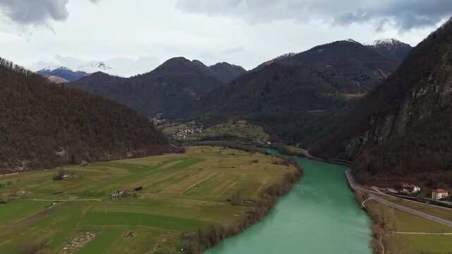 Most na Soči, Slovenia, with the emerald Soča River winding through a forested alpine valley beneath snowy Julian Alps peaks, captured by drone above rural fields and roads.