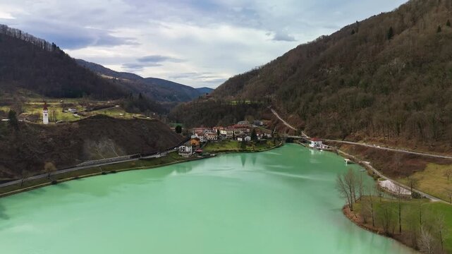 Most na Soči, Slovenia mountain village beside the emerald Soča River and lake in the Julian Alps, with forested winter landscape and village church, captured in aerial view.