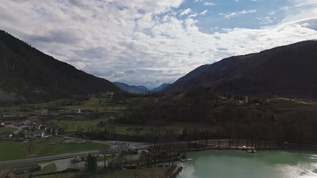 Most na Soči in Slovenia with village homes, church and bridge beside the Soča River and lake in the Julian Alps valley, surrounded by forested hills and winter scenery, drone view.
