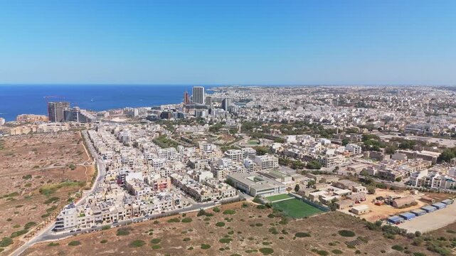 Pembroke, Malta cityscape with Portomaso Tower and Mercury Tower above the Mediterranean coastline, urban residential and business districts with football pitch in aerial view.
