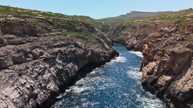 Wied il-Għasri in Gozo, Malta, with a narrow rocky gorge opening to the Mediterranean Sea, clear blue water and rugged cliffs in a tranquil coastal landscape, aerial view.
