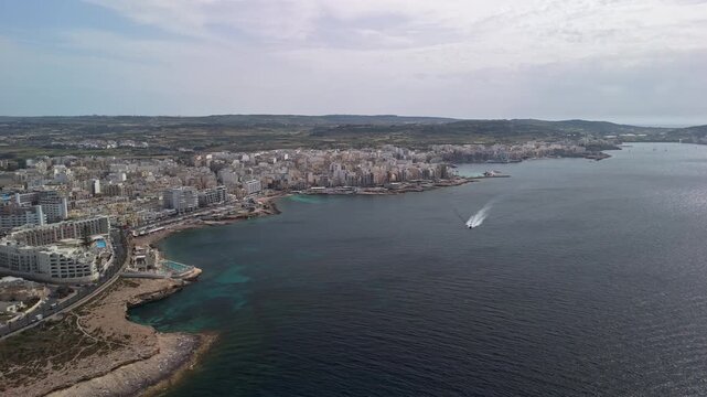 St Paul's Bay and Buġibba waterfront with marina, hotels and residential buildings along the rocky Mediterranean coast and turquoise water, panoramic cityscape captured by drone.
