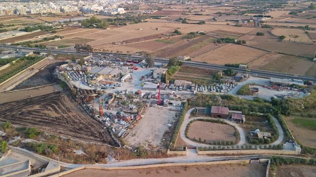Attard, Malta scrapyard and metal recycling center with trucks, cranes and heavy machinery beside fields and warehouses, viewed from above by drone.