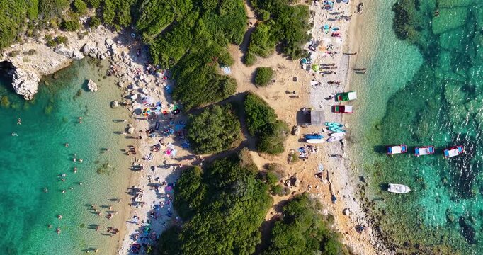 Top-down aerial drone shot of tourists on Porto Timoni twin beach in Afionas, Corfu, Greece. Colorful umbrellas, turquoise water, and boats.