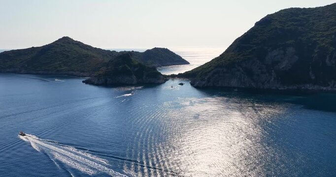 Aerial wide shot of a speedboat cruising near Porto Timoni bay in Corfu, Greece. Sparkling turquoise Mediterranean sea and rocky green coast.
