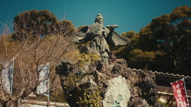 A bronze statue of a winged tengu, the legendary guardian spirit of Hansobo Shrine, perched on a rocky outcrop within the Kencho-ji temple complex in Kamakura, with ceremonial banners visible nearby.