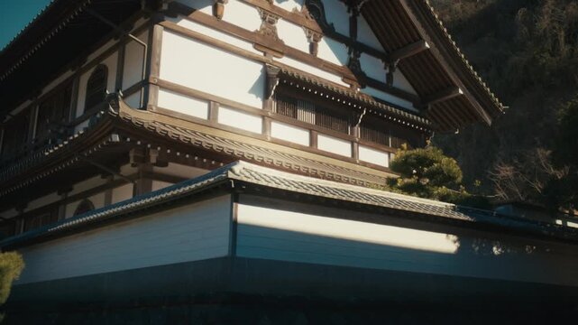 A low-angle view of a traditional two-story temple building with white walls and ornate wooden brackets within the historic Kencho-ji complex in Kamakura, set against a bright sky and hillside greener