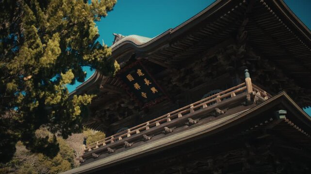 A view of the Sanmon gate's upper story at Kencho-ji temple in Kamakura, showing its inscribed plaque and detailed wooden brackets partially framed by a juniper tree, against a bright blue sky.
