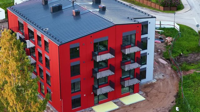 Modern red apartment building with balconies under construction next to a street and green lawn