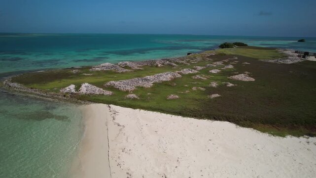 Drone tracking shot of a remote Caribbean island with queen conch shell mounds, white sand beach, and crystal-clear turquoise sea in Los Roques.