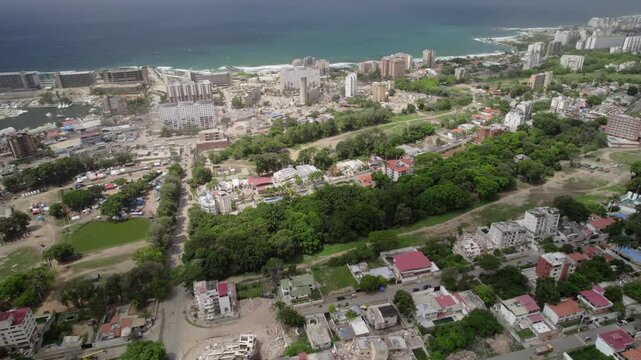 Drone Collapsed Residential Complexes near shore, multiple high-rise buildings are reduced to rubble and dust. Green strip of local golf course stands out clearly, serve temporary shelter camp