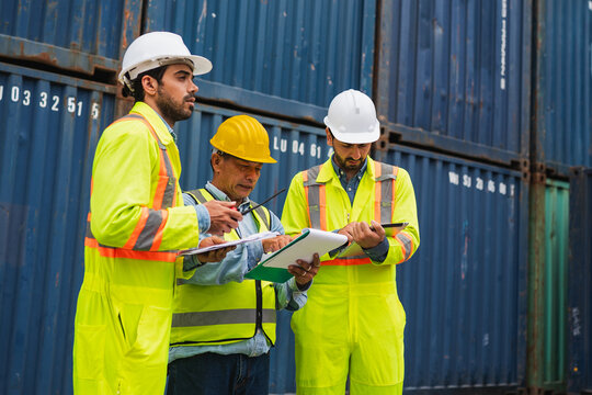 Industrial worker safety vest hard hat clipboard shipping container cargo port inspection logistics teamwork, focused crew review paperwork near stacked blue containers during freight operation