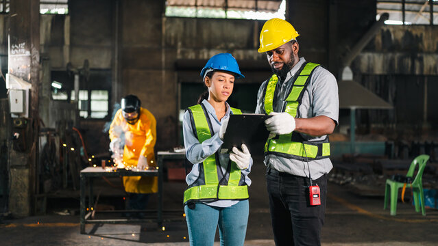 Industrial worker safety vest hard hat clipboard factory inspection with diverse team in metal workshop welding spark protective gear focused supervisor