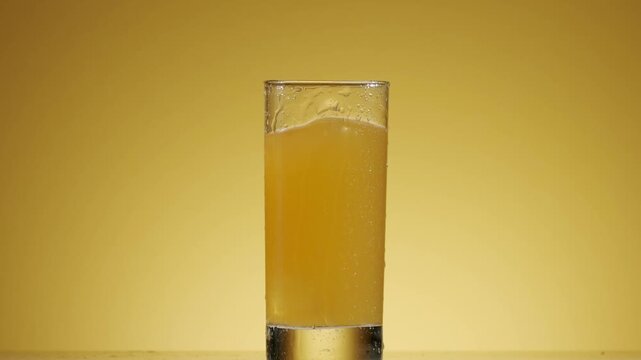 Freshly squeezed orange juice in a tall glass surrounded by whole and sliced ripe oranges on a shiny surface, Refreshing ice cube falling into a tall glass of freshly squeezed orange juice,