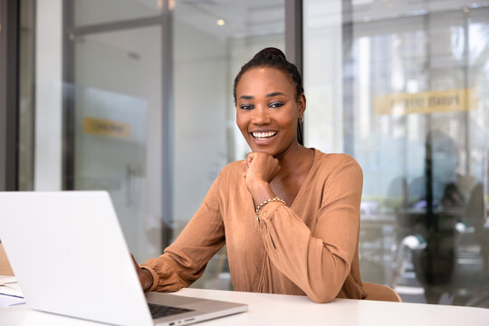 Portrait of happy African professional woman sitting at office desk with laptop, laughs, looks at camera, enjoy workflow using modern device at corporate workplace. Success, career, good business news