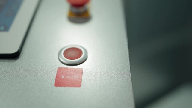 Industrial equipment operator's finger pressing a red stop button on a machine control panel to halt production, with the emergency stop button visible in the background for safety