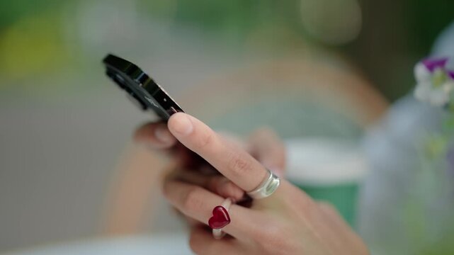 Slow motion close-up of a young woman's stylish hands, with a red heart ring and silver band, using a mobile phone to text, scroll, and browse social media in a bright, modern cafe or space