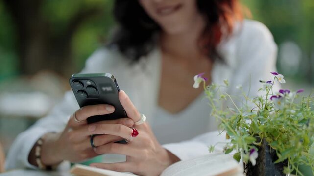 Close-up of a young woman's hands scrolling and texting on her modern smartphone screen while sitting at a table in a garden, surrounded by plants and an open book during a relaxing afternoon