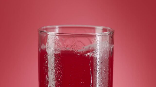 Close up slow motion footage of ice cubes swirling in a refreshing cold red fruit juice inside a wet glass covered with condensation droplets, showing a vibrant and thirst-quenching scene.