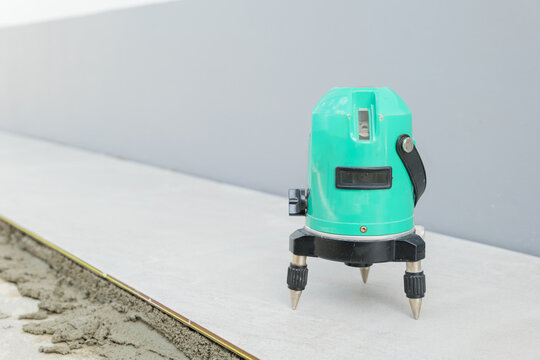 A green laser level on a tripod projects beams across a white and gray floor, ensuring precision while laying ceramic tiles in a construction setting