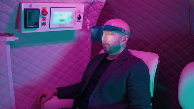 Bald man with a beard sitting inside a hyperbaric chamber during an oxygen therapy session, wearing a headset and breathing through a nasal cannula while relaxing with his eyes closed