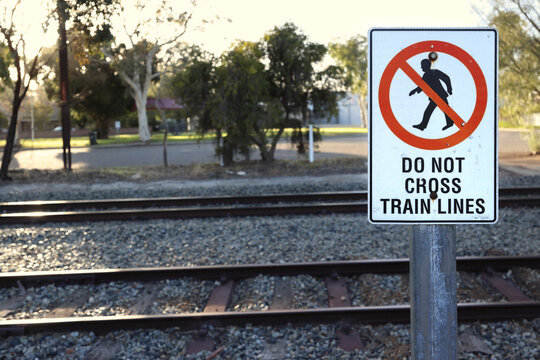 Do not cross train lines warning sign with a pedestrian prohibition symbol posted next to dual railway track in suburban town setting during warm afternoon sunlight