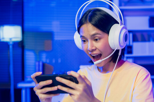 Excited young Asian girl playing mobile game on smartphone and headset at home.