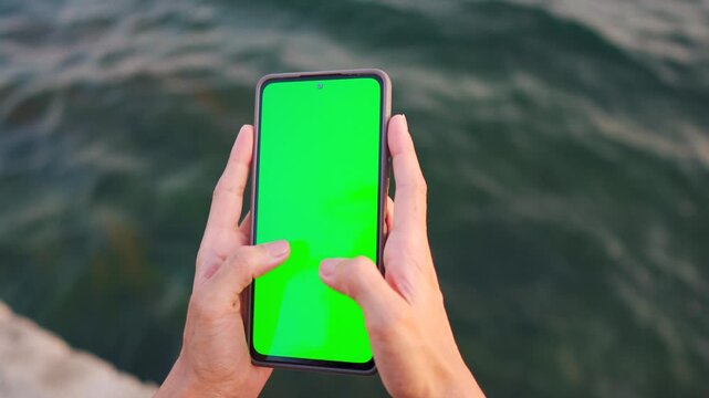 4K Closeup shot of girl hand typing on mobile phone with green screen against lake background. Girl hold mobile closeup. Chroma key page. Wireless technologies, online learning and shopping. 