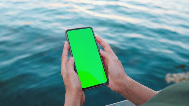 4K Shot of Woman's hand holding smartphone with green mock-up screen in vertical mode. Mobile phone green screen against water background. Chroma key mock up for advertising, social networks. 