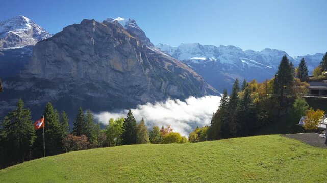 Murren autumn alpine landscape, Swiss flag, green meadow, sea of clouds and Jungfrau mountain peak. Murren, Canton of Bern, Switzerland.