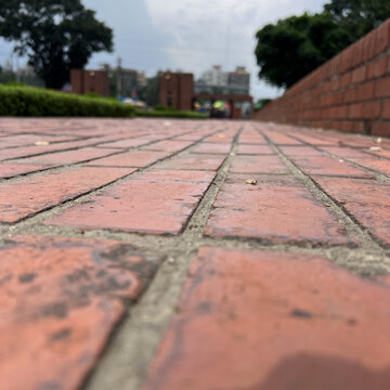 A low angle perspective of a red brick paved pathway leading towards a blurred park background, with trees and, brick structures under, a cloudy sky