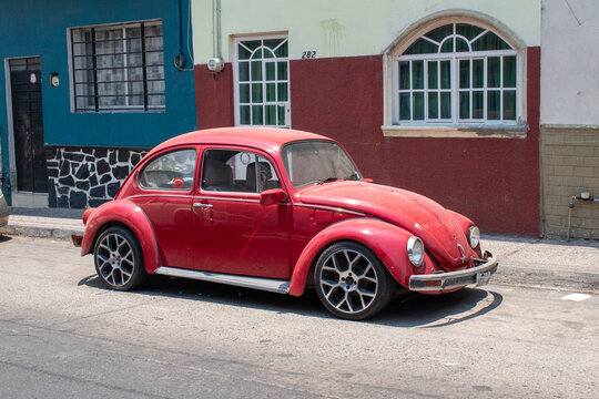 Guadalajara, Mexico - May 17 2025: Old red VW Beetle with modified rims parked in front of colorful houses in Guadalajara