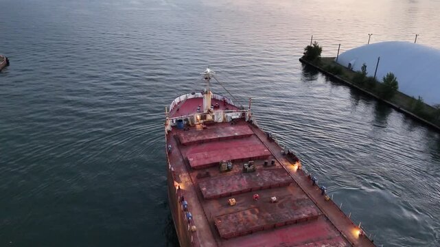 High-angle stationary shot looking down at the red and weathered steel deck and bow of a massive cargo ship navigating slowly past a white storage dome building in a port channel in Toronto