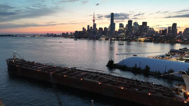 High-angle stationary shot looking down at the long steel deck, hatches, and self-unloading boom of a bulk carrier cargo ship navigating past the illuminated Toronto skyline at dusk