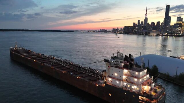 High-angle stationary shot of a massive industrial bulk carrier ship moving slowly down a port channel, past an illuminated storage dome with the glowing Toronto skyline at dusk