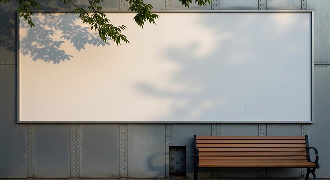 A large empty rectangular notice board hangs upon a textured industrial wall surface near a wooden bench while dappled tree foliage shadows cast a natural pattern across the broad vacant signage