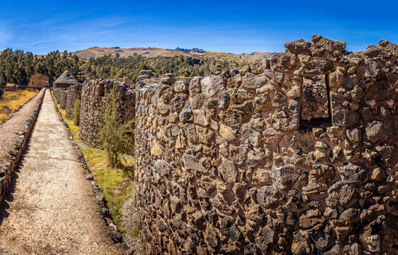 Ancient stone pathway leading to traditional buildings at the archaeological site of Raqchi surrounded by Andean highland scenery under a clear blue sky