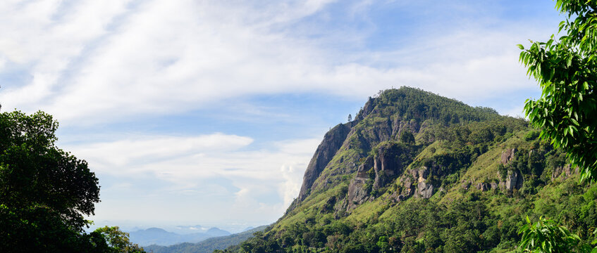 Green mountain landscape with blue sky, rugged rocky cliffs, forested slopes, in Sri Lanka's hill country.