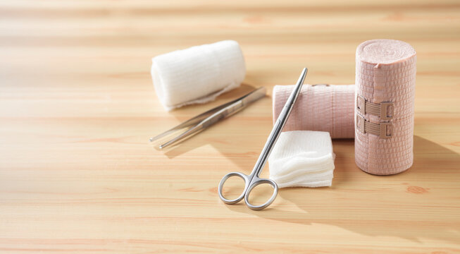 Medical first aid supplies including elastic bandages, sterile gauze pads, and surgical scissors arranged on a wooden table. Healthcare, emergency treatment, injury care