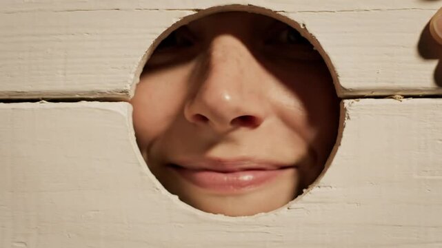 Close up shot of a person's face framed within a round hole in a white wooden stockade, smiling