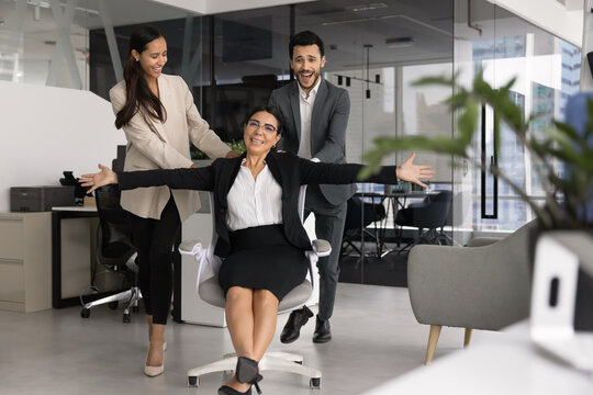 Happy diverse business team celebrating corporate success, two colleagues playfully pushing smiling female coworker sits on office chair, riding, laughing, having fun during workday break at workplace