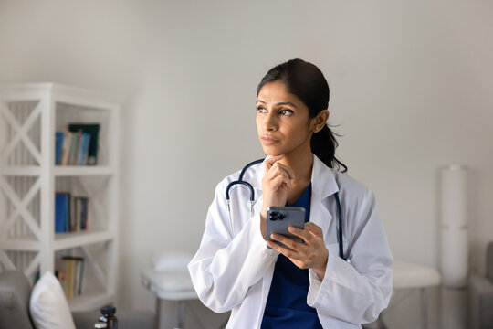 Thoughtful South Asian doctor standing in office, holding smartphone, thinking on treatment or diagnosis, analyzing data, researching information in clinic. Digital communication, medical app use
