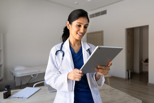 Adult South Asian female doctor reviewing patient data on digital tablet, working on treatment plan or researching information in clinic office. Telehealth, medical technology, healthcare services
