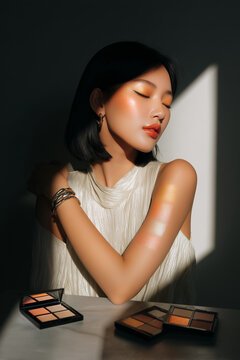 Beauty portrait of a woman with black bob hair, glowing makeup and shimmer swatches on her arm in dramatic studio light showcasing modern cosmetics and glamour