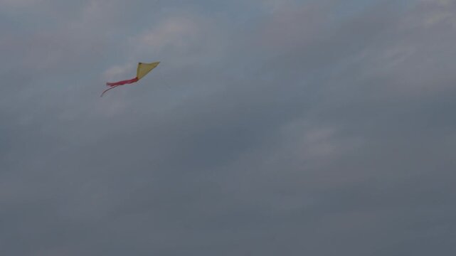 Single kite soars above a cloudy sky. Its yellow body glows gently against the gray. Red tail streams behind it as it dances. Wind carries it effortlessly, free and light. Ideal for calm, joyful