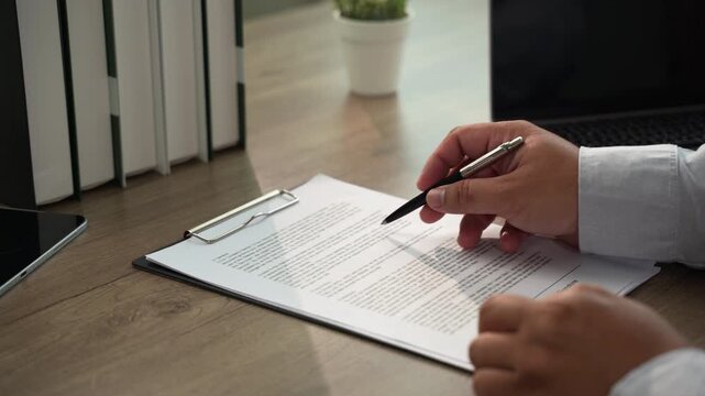 Hand signs a printed contract with pen on clipboard at a wooden desk beside laptop, using daylight workspace for agreement signing, paperwork handling, and contract documentation workflow.