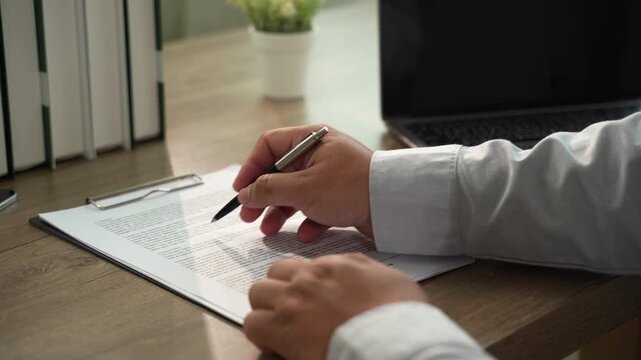 Hand signs a contract document with pen on clipboard at desk beside laptop, while another hand points to text for agreement approval, paperwork review, and signing workflow in daylight.
