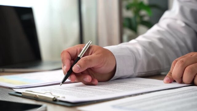 Hand signs a contract document with pen on clipboard at desk beside laptop, while another hand points to text for agreement approval, paperwork review, and signing workflow in daylight.